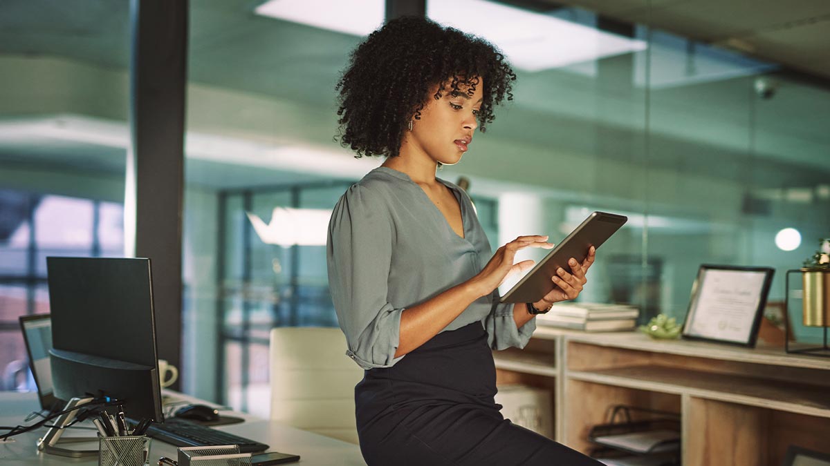 A woman leaning against a desk in an office while using a tablet