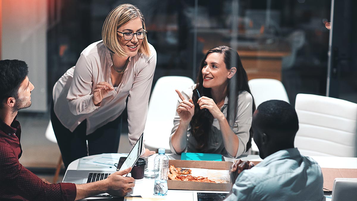 A group of colleagues sat around a table in the office, laughing and eating pizza