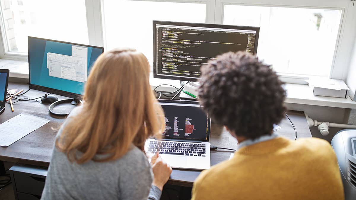 Two people at a desk with a laptop and monitors looking at the screens 