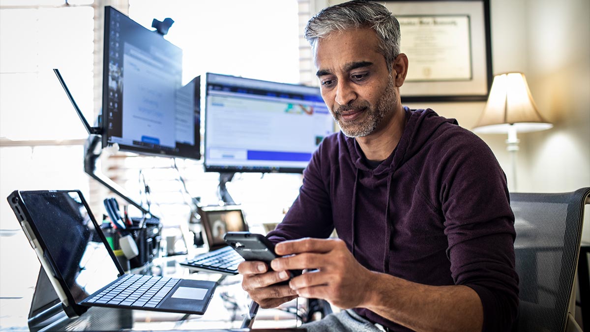 A middle aged man sat on his phone in his home office, next to two monitor screens