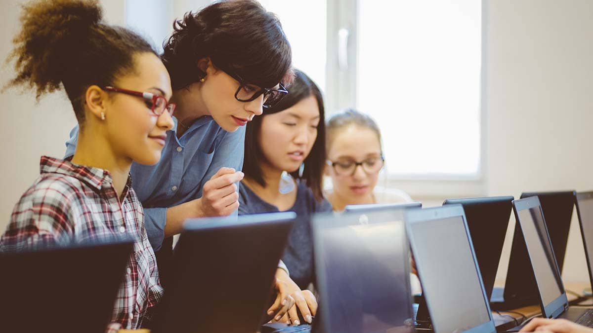 Three female students with a teacher in a school environment working on laptops.