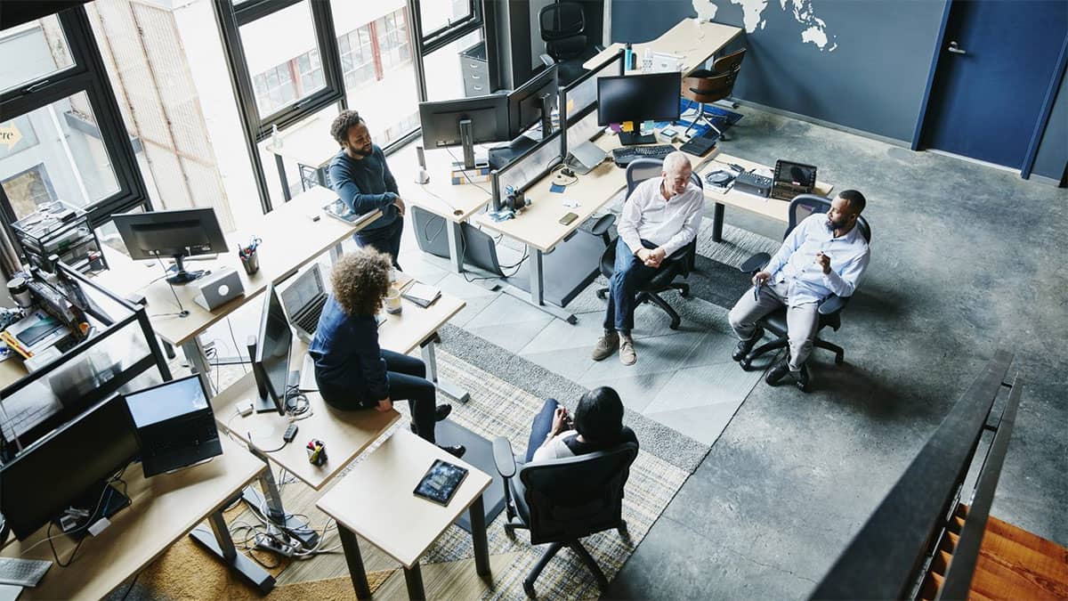 Five people in an office space having an informal meeting, dressed in casual business attire.